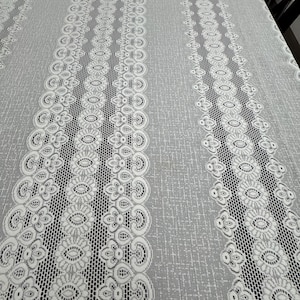 May include: A rectangular tablecloth featuring a light gray base with a subtle textured pattern. The fabric is decorated with vertical bands of white lace that display intricate floral and scrollwork motifs, separated by sections of openwork mesh.