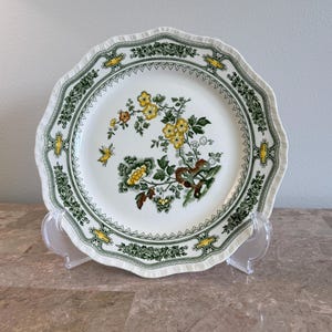 May include: A white ceramic plate with a scalloped edge featuring a central botanical illustration of yellow flowers, green leaves, and a small butterfly. The rim is decorated with a dark green geometric border and yellow accents.