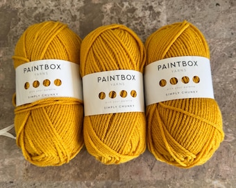 Puede incluir: Tres ovillos de lana color amarillo mostaza, cada uno envuelto en una etiqueta de papel blanco. Las etiquetas muestran el texto "PAINTBOX YARNS" y "SIMPLY CHUNKY" con cuatro recortes circulares que dejan ver el color.