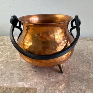 May include: A round, hammered copper pot with a wide opening and a polished finish. The pot features a thick, black, curved metal handle attached to the sides and stands on three small, tapered metal legs that provide a stable base.