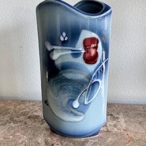 May include: A ceramic vase featuring a gradient of blue tones, ranging from deep navy to light sky blue. The surface is decorated with an abstract white and dark red graphic pattern, including circular and linear shapes. The vase has a curved rim.