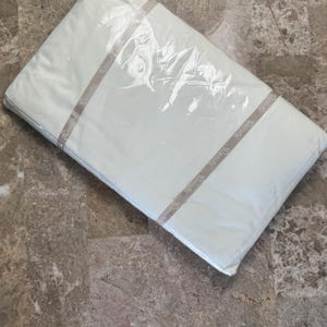 Puede incluir: Un paquete rectangular que contiene tela blanca, envuelto en pl&aacute;stico transparente. Una banda de papel beige envuelve el centro del paquete, con el nombre de la marca Wamsutta impreso repetidamente en una fuente oscura a lo largo de la banda.
