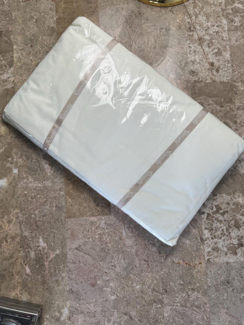 Puede incluir: Un paquete rectangular que contiene tela blanca, envuelto en pl&aacute;stico transparente. Una banda de papel beige envuelve el centro del paquete, con el nombre de la marca Wamsutta impreso repetidamente en una fuente oscura a lo largo de la banda.