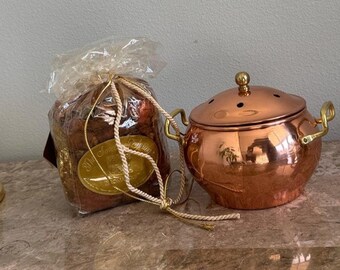 Puede incluir: Una bolsa de plástico transparente llena de popurrí marrón con una etiqueta ovalada dorada que dice The Sweet Spring, junto a una olla de cobre pulido con tapa a juego, dos asas laterales y un pequeño pomo dorado.