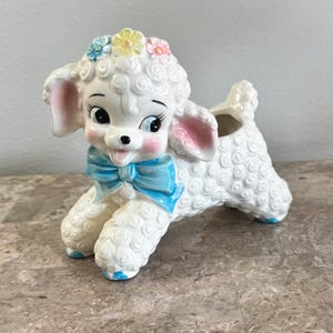 May include: A white ceramic lamb planter featuring a textured curly wool finish, a bright blue bow around its neck, and pastel-colored flowers on its head. The lamb has pink-tinted ears, large dark eyes, and blue accents on its hooves.