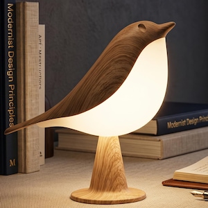 Puede incluir: Una lámpara de mesa con forma de pájaro que presenta una base y un dorso con textura de veta de madera, junto con un pecho blanco suave que difunde la luz. La lámpara se apoya en un pedestal circular y emite una iluminación cálida.