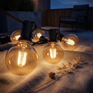 May include: A string of outdoor globe lights featuring clear glass bulbs with visible warm-toned LED filaments. The bulbs are connected by a black cord and emit a soft, golden glow, resting on a textured fabric surface with a dried plant stem.