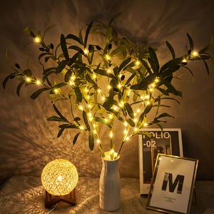 May include: An artificial olive branch arrangement with green leaves and small black olives, illuminated by integrated warm white LED string lights, displayed in a white ceramic vase alongside a spherical woven lamp and a framed letter M.