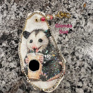 May include: A decorative oyster shell ornament featuring a painted opossum holding a coffee cup, embellished with a rhinestone border and a gold-tone lobster clasp with pink and white beads. The text Glenda Sue is printed in pink on the shell.