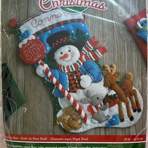 May include: A Bucilla felt Christmas stocking craft kit featuring a snowman, reindeer, squirrel, and mouse. The stocking includes a red sign that reads Santa Please Stop Here, a candy cane, and measures 18 inches. It includes felt, floss, sequins, beads, and needles.