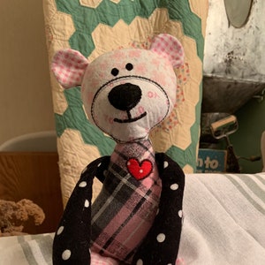 May include: A handmade patchwork teddy bear with pink and black fabric. The bear has a black nose and eyes, and a red heart on its chest. The bear is sitting on a white and gray striped towel.