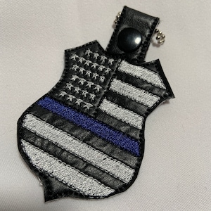 May include: A black leather keychain shaped like a police badge with an embroidered American flag and a blue line. The keychain has a silver chain and a black button.