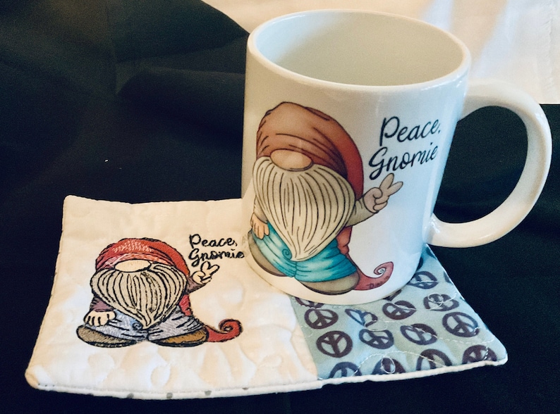 May include: A white ceramic mug with a gnome design and the text "Peace, Gnomie". The mug is sitting on a white and blue fabric coaster with a gnome design and peace signs.