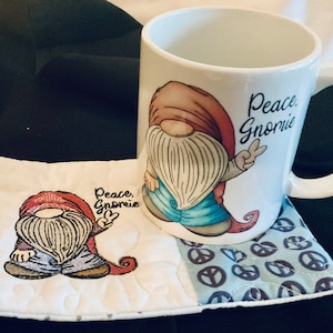 May include: White ceramic mug with a gnome design and the text "Peace, Gnomie". The mug is sitting on a white and blue quilted coaster with a peace sign design.