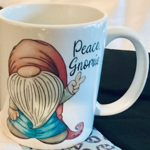May include: White ceramic coffee mug with a cartoon gnome wearing a red hat, blue jeans, and a white beard. The gnome is holding up two fingers in a peace sign. The text "Peace Gnome" is printed on the mug.