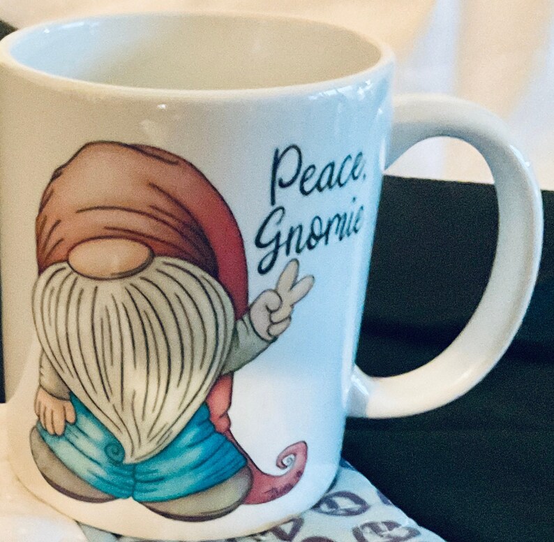 May include: White ceramic coffee mug with a cartoon gnome wearing a red hat, blue jeans, and a white beard. The gnome is holding up two fingers in a peace sign. The text "Peace Gnome" is printed on the mug.