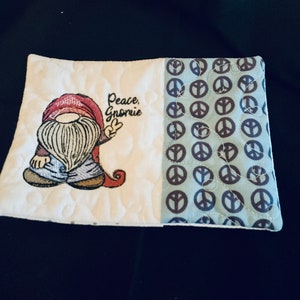 May include: A white quilted hot pad with a gnome holding a peace sign and the text "Peace, Gnome" on one side and a blue fabric with brown peace signs on the other side.