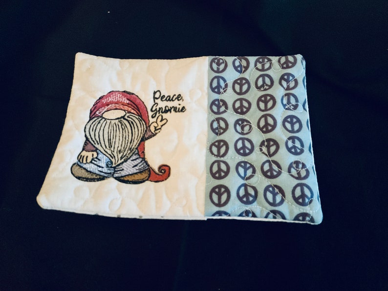 May include: A white quilted hot pad with a gnome holding a peace sign and the text "Peace, Gnome" on one side and a blue fabric with brown peace signs on the other side.