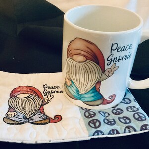 May include: A white ceramic mug with a gnome design and the text "Peace, Gnomie". The mug is sitting on a white and blue quilted coaster with a peace sign pattern.