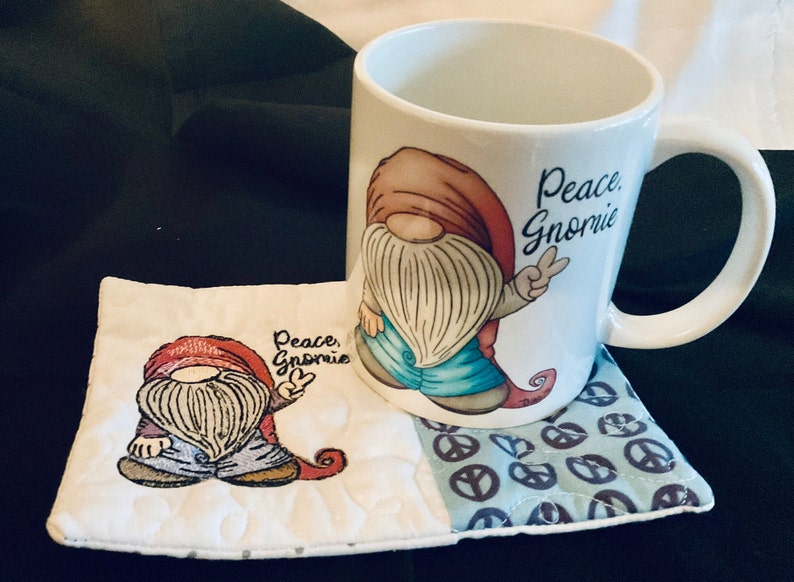 May include: A white ceramic mug with a gnome design and the text "Peace, Gnomie". The mug is sitting on a white and blue quilted coaster with a peace sign pattern.