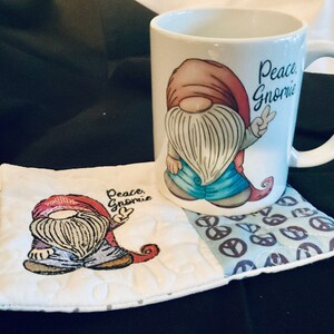 May include: White ceramic mug with a red and blue gnome design and the text "Peace Gnomie". The mug is sitting on a white and blue quilted coaster with a peace sign pattern.