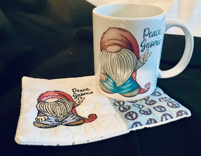 May include: White ceramic mug with a red and blue gnome design and the text "Peace Gnomie". The mug is sitting on a white and blue quilted coaster with a peace sign pattern.
