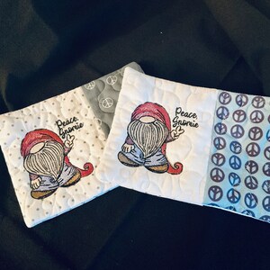 May include: Two white fabric coasters with a gnome design and the text "Peace, Gnome" embroidered on them. The coasters are made of a patchwork of fabrics with a peace sign pattern on one side.