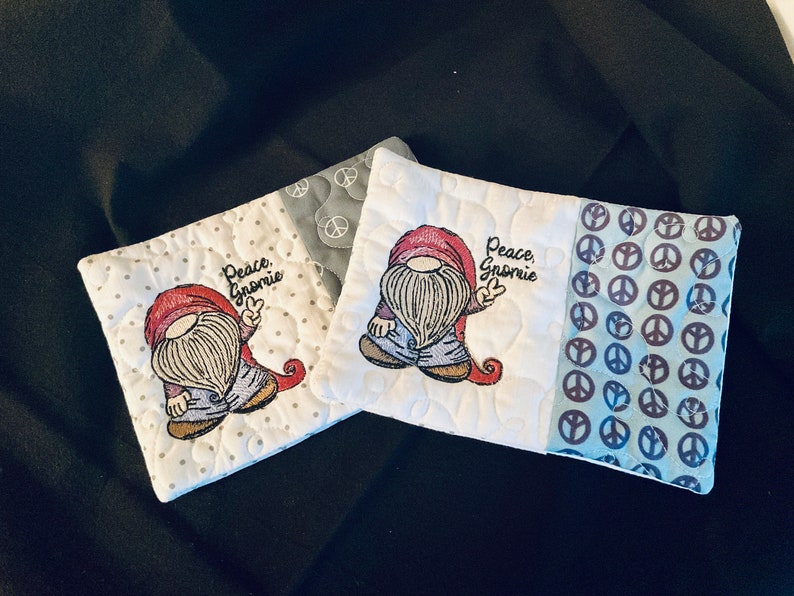 May include: Two white fabric coasters with a gnome design and the text "Peace, Gnome" embroidered on them. The coasters are made of a patchwork of fabrics with a peace sign pattern on one side.
