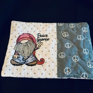 May include: A white and gray quilted coaster with a gnome embroidered on it. The gnome is wearing a red hat, blue pants, and a white beard. The text "Peace, Gnome" is embroidered above the gnome. The other side of the coaster is gray with a white peace sign pattern.