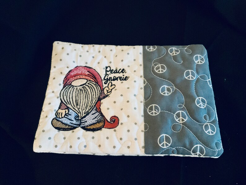May include: A white and gray quilted coaster with a gnome embroidered on it. The gnome is wearing a red hat, blue pants, and a white beard. The text "Peace, Gnome" is embroidered above the gnome. The other side of the coaster is gray with a white peace sign pattern.