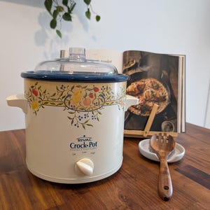 Peut inclure: Une mijoteuse Rival Crock-Pot de couleur crème avec un bord bleu foncé et une bordure à motifs de fruits représentant des poires, des baies et des feuilles. Elle possède un couvercle en verre transparent, deux poignées latérales et un bouton.