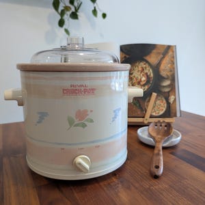 May include: A Rival Crock-Pot stoneware slow cooker with a cream-colored exterior, featuring horizontal pastel pink and blue stripes and a central floral motif. It includes a clear glass lid, two side handles, and a front dial with Off, Low, and High settings.
