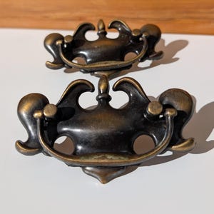 May include: Two matching drawer pulls made of dark bronze-toned metal. Each pull features a decorative backplate with curved, symmetrical scrollwork and a central finial, paired with a hinged, semi-circular drop handle attached by two posts.
