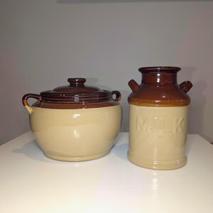 Peut inclure: Un ensemble en céramique composé d'un pot rond avec couvercle et deux anses latérales, ainsi qu'un pichet à lait cylindrique avec deux petites anses. Les deux pièces présentent un design bicolore avec une base crème et un haut brun foncé.