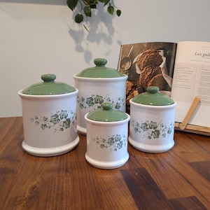 Peut inclure: Un ensemble de quatre boîtes de conservation en céramique blanche de tailles variées, ornées d'un motif de feuilles de lierre vertes et dotées d'un couvercle vert assorti. Elles sont cylindriques avec une base évasée sur une table en bois.