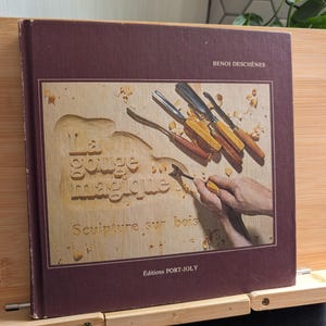Peut inclure: Un livre à couverture rigide carrée de couleur brun foncé intitulé La gouge magique Sculpture sur bois par Benoi Deschênes. La couverture présente une image d'outils de sculpture sur bois et une main utilisant une gouge sur du bois.