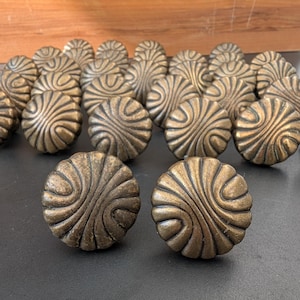 May include: A collection of round, antique brass-toned metal cabinet knobs. Each knob features a raised, swirling, fan-like pattern radiating from the centre to the outer edge, creating a textured, circular design with a dark, weathered finish.