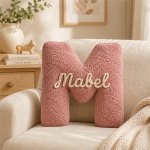 Pode incluir: Uma almofada em forma de letra M na cor rosa seco com um acabamento texturizado tipo sherpa. O nome Mabel está bordado em letra cursiva de cor creme no centro da letra, criando uma peça decorativa suave e tátil.