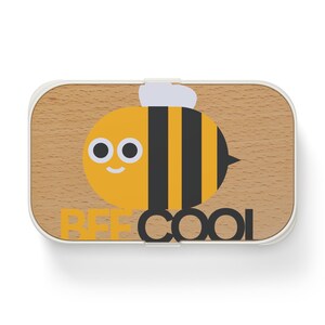 Peut inclure: Une boîte à déjeuner rectangulaire avec un couvercle au fini grain de bois clair. Le couvercle présente un graphique d'une abeille rayée jaune et noire avec des ailes blanches et le texte BEE COOL imprimé en lettres jaunes et noires en bas.