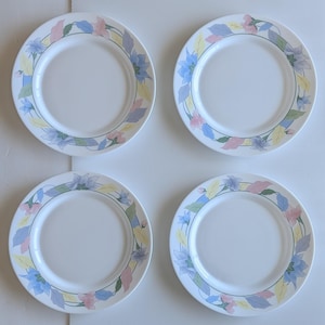 May include: A set of four round white ceramic plates, each featuring a decorative border of pastel-coloured floral illustrations. The pattern consists of stylised blue, pink, and yellow leaves and blossoms arranged in a continuous ring around the rim.