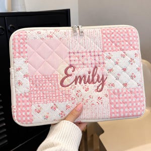 May include: A rectangular quilted laptop sleeve featuring a patchwork design of pink and white gingham, floral patterns, and solid pink squares. The name Emily is embroidered in dark pink script in the center. It has a white zipper closure.