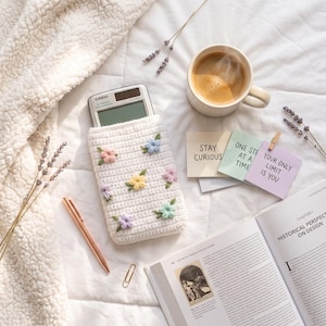 Peut inclure: Une pochette crochetée blanche ornée de petites fleurs pastel, contenant une calculatrice Casio. La pochette est entourée d'une tasse de café en céramique, d'un stylo or rose, d'un trombone et de trois notes adhésives avec du texte.