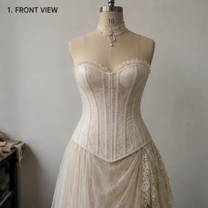 May include: A cream-colored strapless corset top featuring a sweetheart neckline with delicate lace trim, vertical boning, and a matching lace choker with a small pendant. The text 1. FRONT VIEW is visible in the upper left corner of the image.