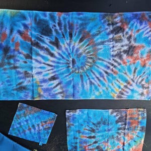 May include: A set of rectangular and square towels featuring a vibrant tie-dye pattern in shades of bright blue, deep purple, black, and hints of orange. The fabric displays a prominent spiral design in the center of the largest towel.