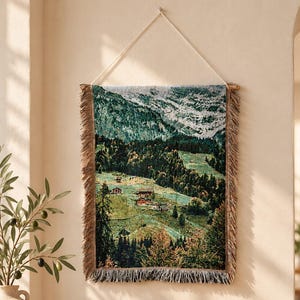 May include: A rectangular woven wall tapestry featuring a detailed landscape of a mountain range, dense green forests, and small houses nestled in a valley. The tapestry has a fringed border on the sides and bottom and hangs from a wooden rod.