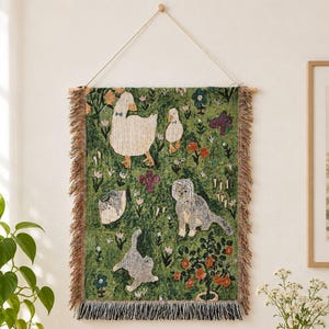 May include: A rectangular woven wall hanging featuring a green background with illustrations of white ducks, a grey cat, and a small grey animal. The tapestry includes orange and blue flowers, a small tree, and decorative fringe along the bottom edge.