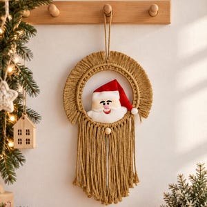 Könnte beinhalten: Ein Makramee-Wandbehang mit einem kreisförmigen Rahmen, beigen Kordelfransen und einem mittigen Plüsch-Weihnachtsmanngesicht mit rot-weißer Mütze. Das Ornament verfügt über eine Aufhängeschlaufe oben und lange, herabhängende Fransen am unteren Ende.
