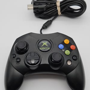 May include: A black wired game controller featuring a central Xbox logo button, two black analog sticks, a directional pad, and four colored action buttons labeled A, B, X, and Y. The controller includes a black cable with a connector plug.