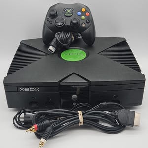 May include: A black original Xbox video game console featuring a prominent green circular logo on the top, accompanied by a black wired controller and a set of black AV cables with red, white, and yellow connectors for audio and video output.