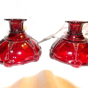 May include: A pair of deep red glass candle holders featuring a wide, rounded base with raised beaded detailing radiating from the center. Each piece has a short, cylindrical neck and a flared rim, showcasing a translucent, glossy finish.